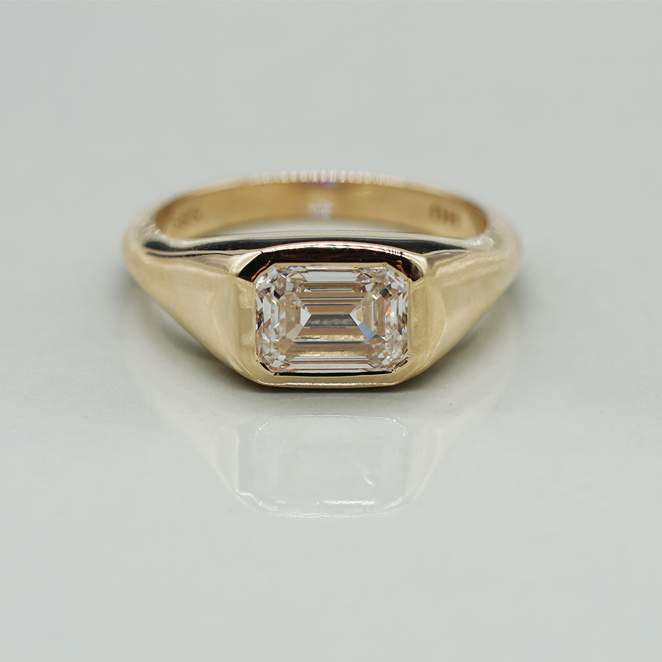 A yellow gold signet ring featuring a rectangular emerald-cut clear gemstone, displayed on a clean, off-white reflective surface.