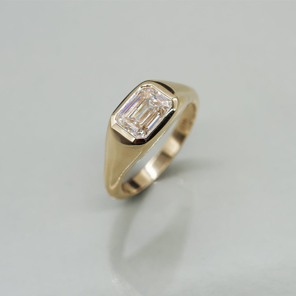 A yellow gold ring featuring an emerald-cut clear diamond, photographed on a clean, light reflective surface.