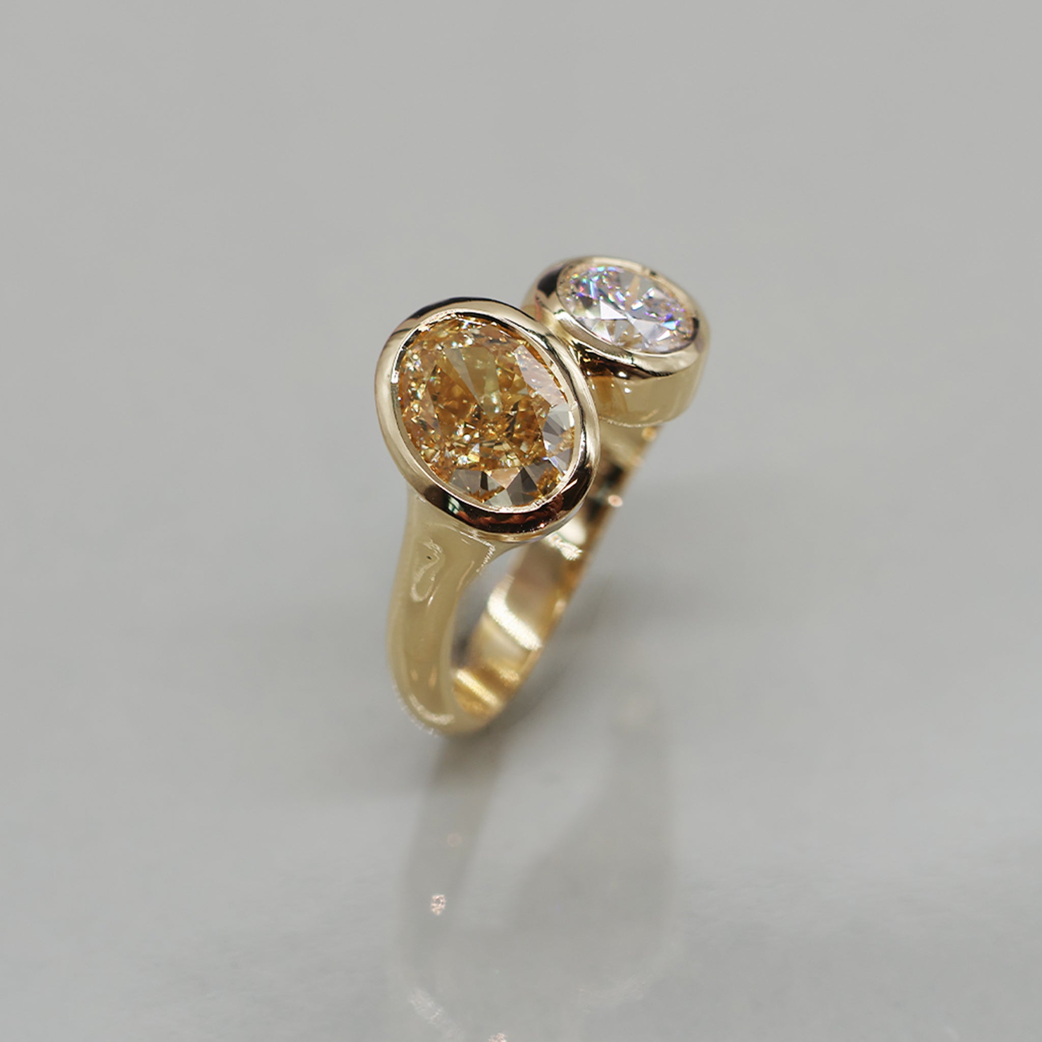 A toi et moi oval ring with two oval-cut gemstones, featuring a prominent warm champagne-colored stone and a smaller clear diamond, set against a neutral light background.