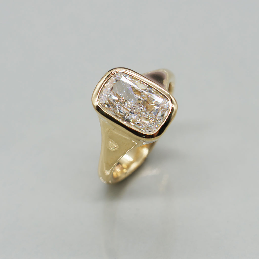 A gold ring featuring a large rectangular cushion-cut clear gemstone in a bezel setting, shown on a neutral light background.