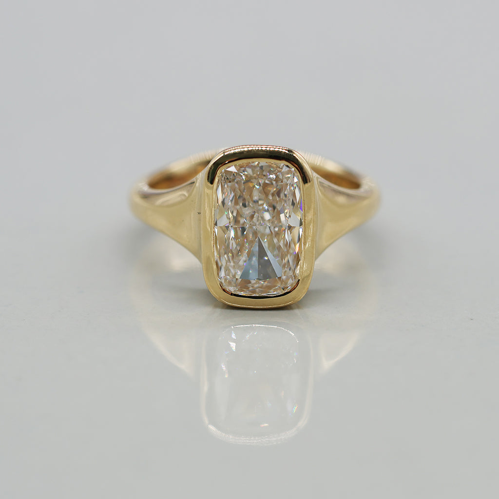 A gold ring featuring a large cushion-cut diamond in a bezel setting, displayed on a clean light reflective surface.