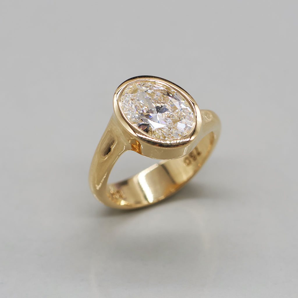 A yellow gold ring featuring a large, round brilliant-cut diamond in an oval bezel setting, displayed on a clean, light reflective surface.