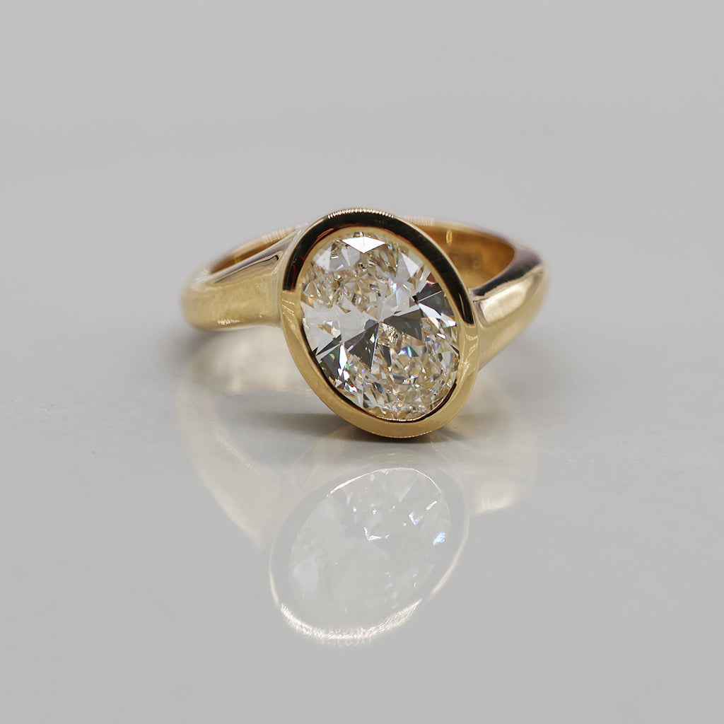 A yellow gold ring featuring a large, round brilliant-cut diamond in an oval bezel setting, displayed on a clean, light reflective surface.