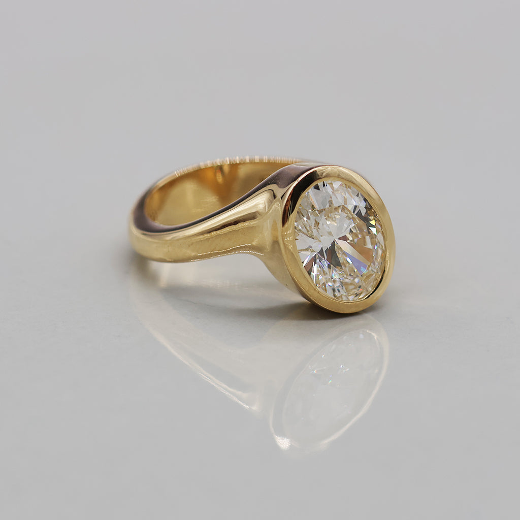A yellow gold ring featuring a large, round brilliant-cut diamond in an oval bezel setting, displayed on a clean, light reflective surface.