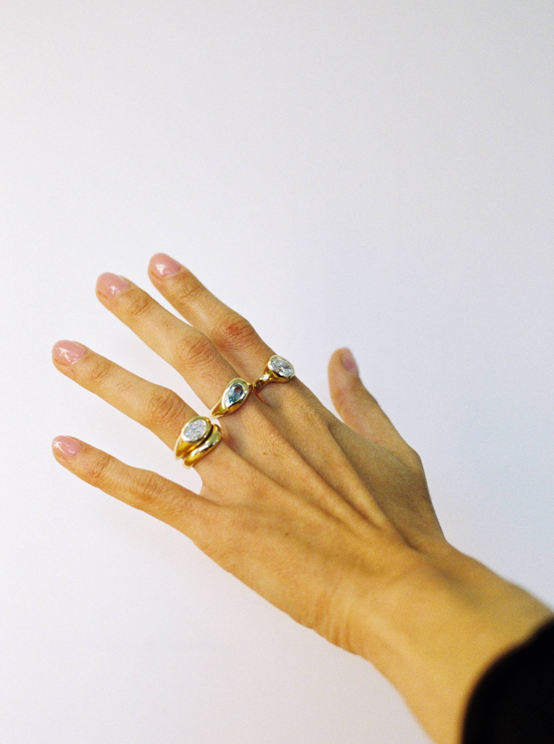 Hand wearing 4 different style gold rings with gemstones and diamonds on a plain background