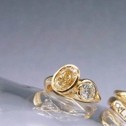 A pair of gold rings with oval and round diamond settings, displayed on a glossy white surface against a dark gray gradient background.
