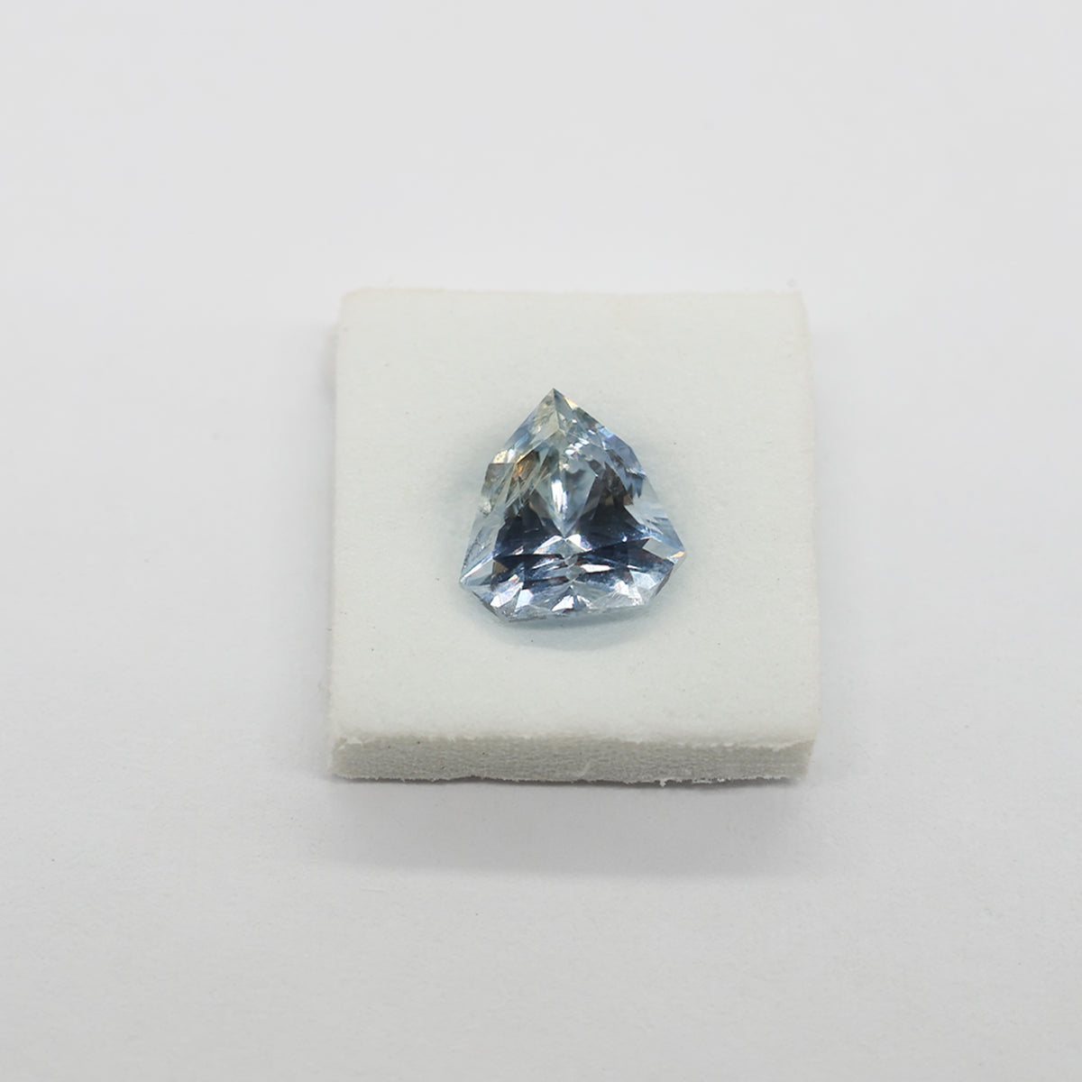 A light blue triangular faceted sapphire displayed on a small white block against a plain light background.
