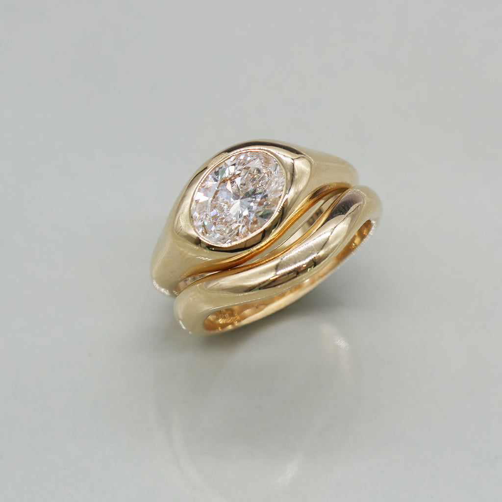A yellow gold oval ring featuring a large oval brilliant-cut diamond center stone paired with matching stacker set against a clean white background with a soft reflection.