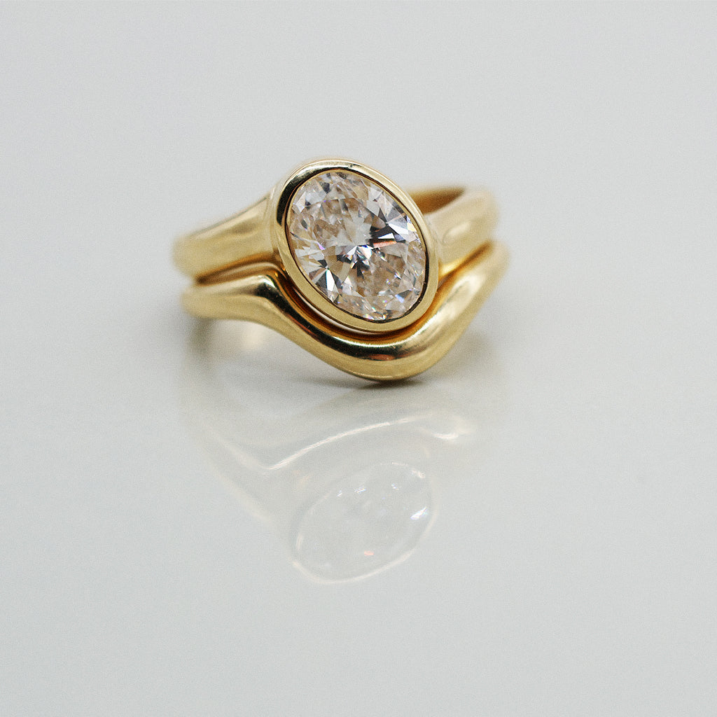 Gold ring asymmetric design with oval diamond and matching paired wedding band, solid gold on a clear reflective white background 