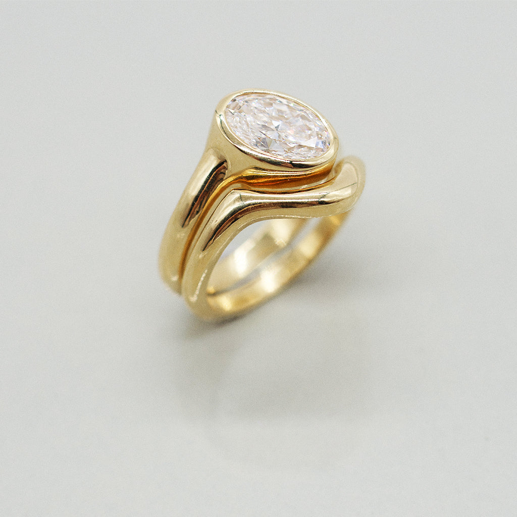 Gold ring asymmetric design with oval diamond and matching paired wedding band, solid gold on a clear reflective white background 