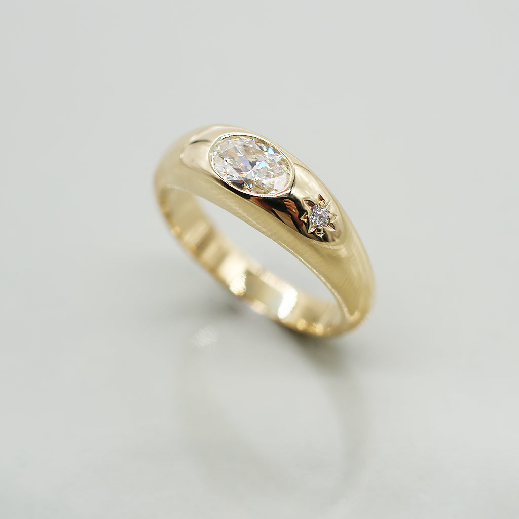 A yellow gold oval ring featuring a large oval brilliant-cut diamond center stone and a small round diamond accent on the band, set against a clean white background with a soft reflection.