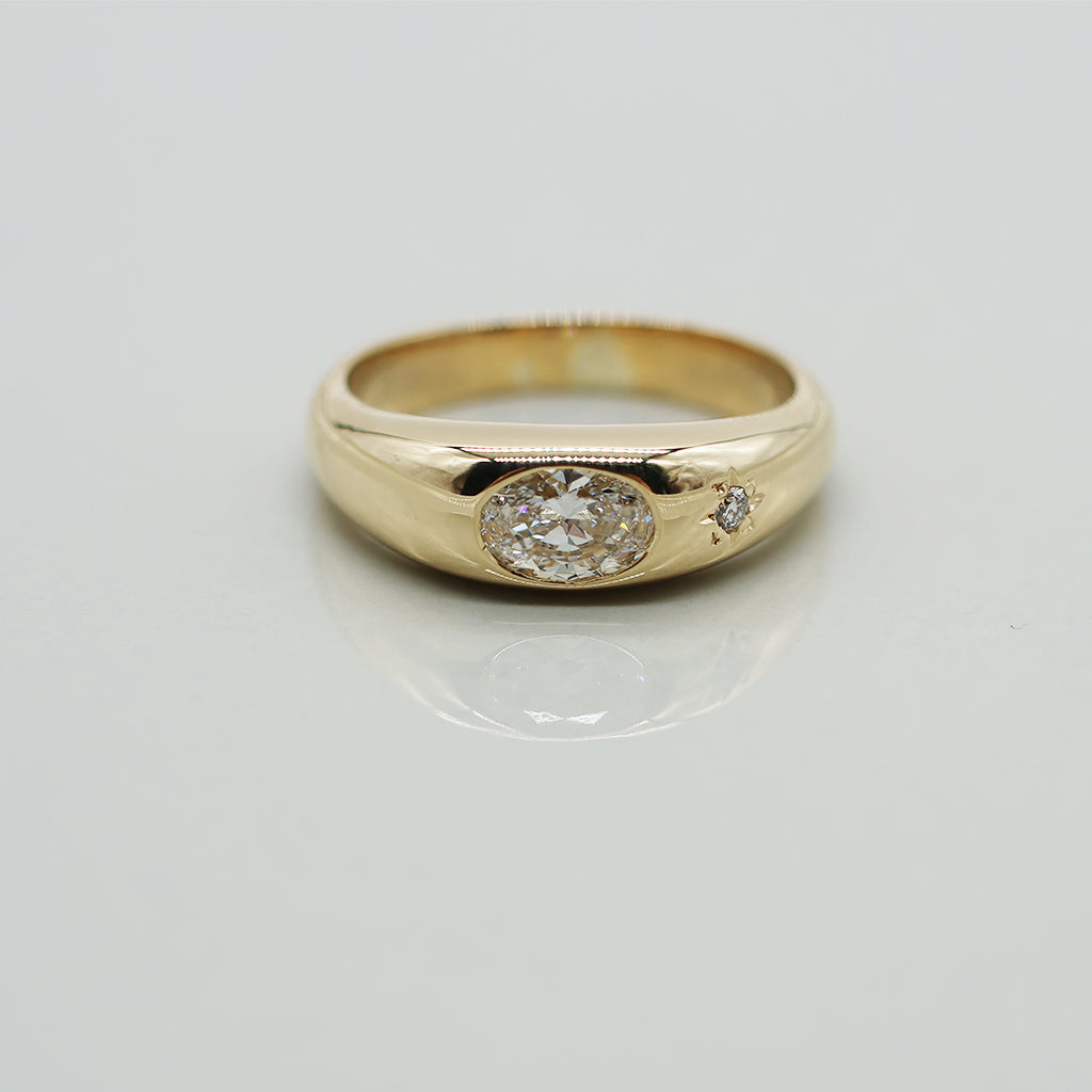 A yellow gold oval ring featuring a large oval brilliant-cut diamond center stone and a small round diamond accent on the band, set against a clean white background with a soft reflection.