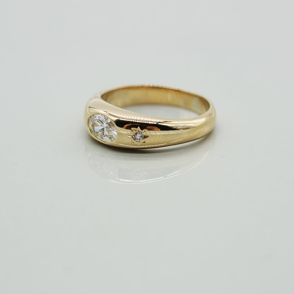 A yellow gold oval ring featuring a large oval brilliant-cut diamond center stone and a small round diamond accent on the band, set against a clean white background with a soft reflection.