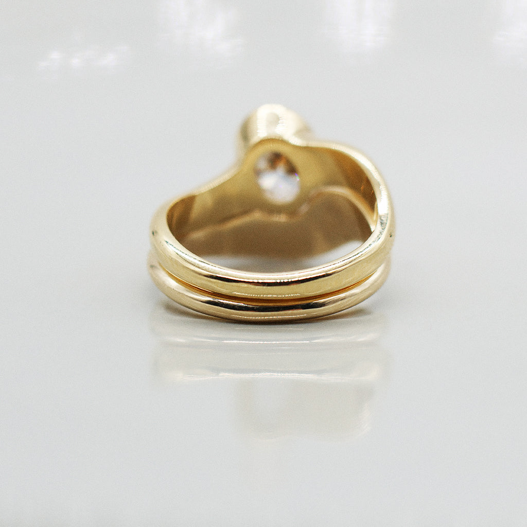 Gold ring asymmetric design with oval diamond and matching paired wedding band, solid gold on a clear reflective white background. 