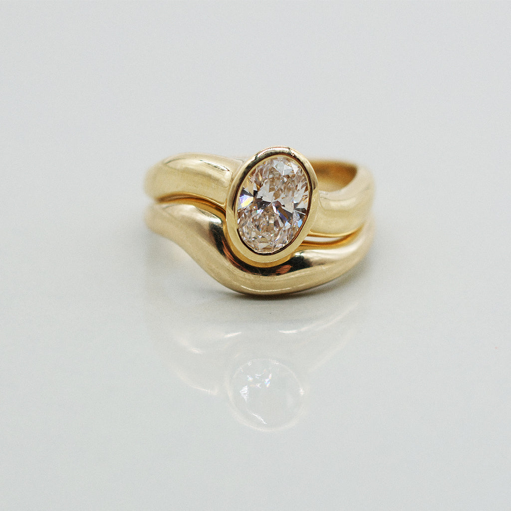 Gold ring asymmetric design with oval diamond and matching paired wedding band, solid gold on a clear reflective white background. 