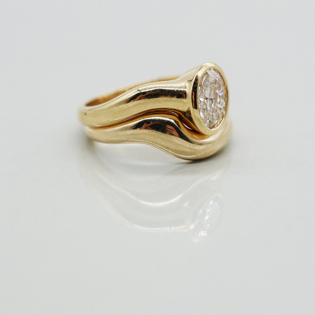 Gold ring asymmetric design with oval diamond and matching paired wedding band, solid gold on a clear reflective white background. 