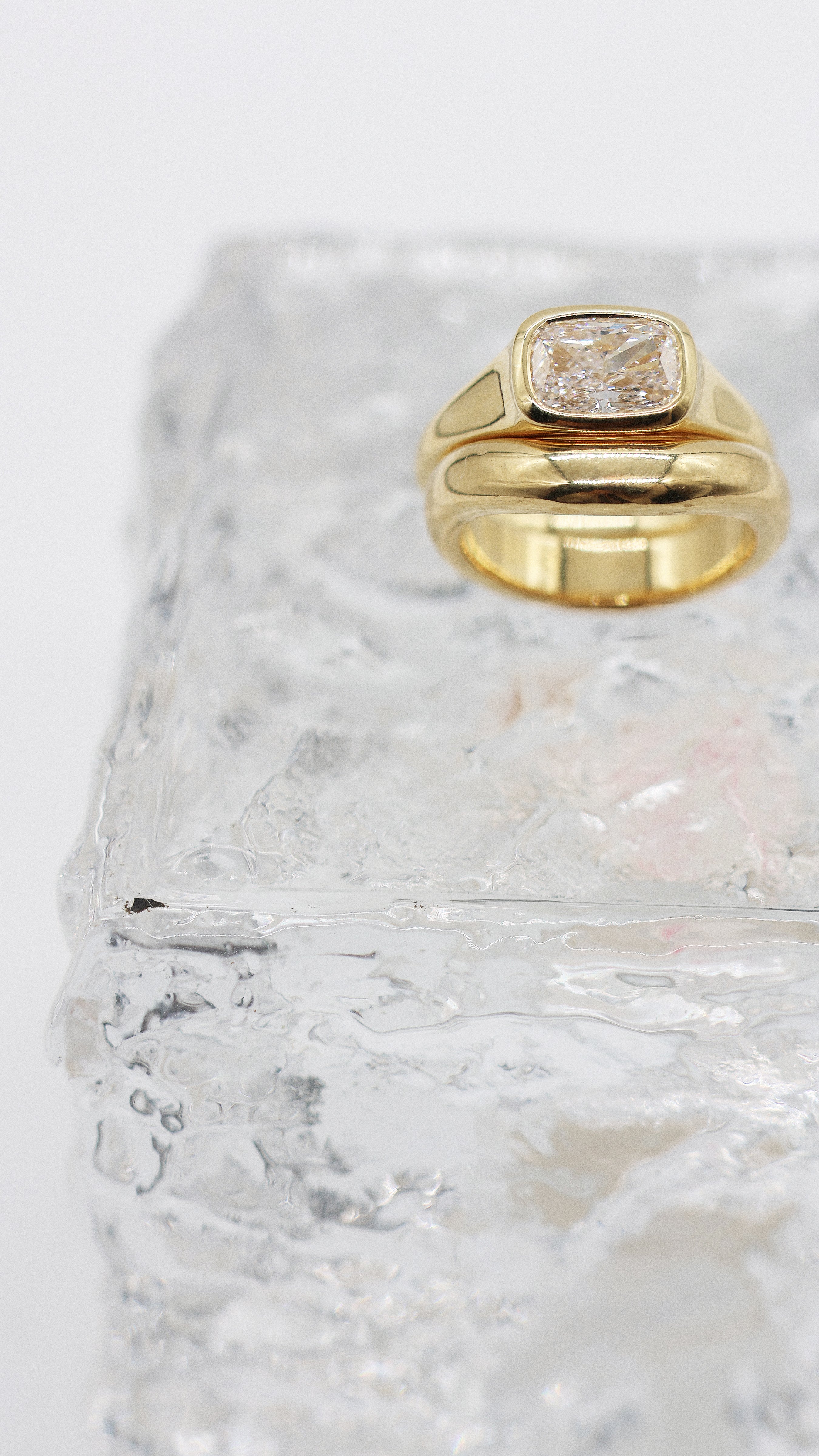A yellow gold cushion ring featuring a large cushion brilliant-cut diamond center stone, paired with a wedding band, set against a clean white background with a glass brick soft reflection.