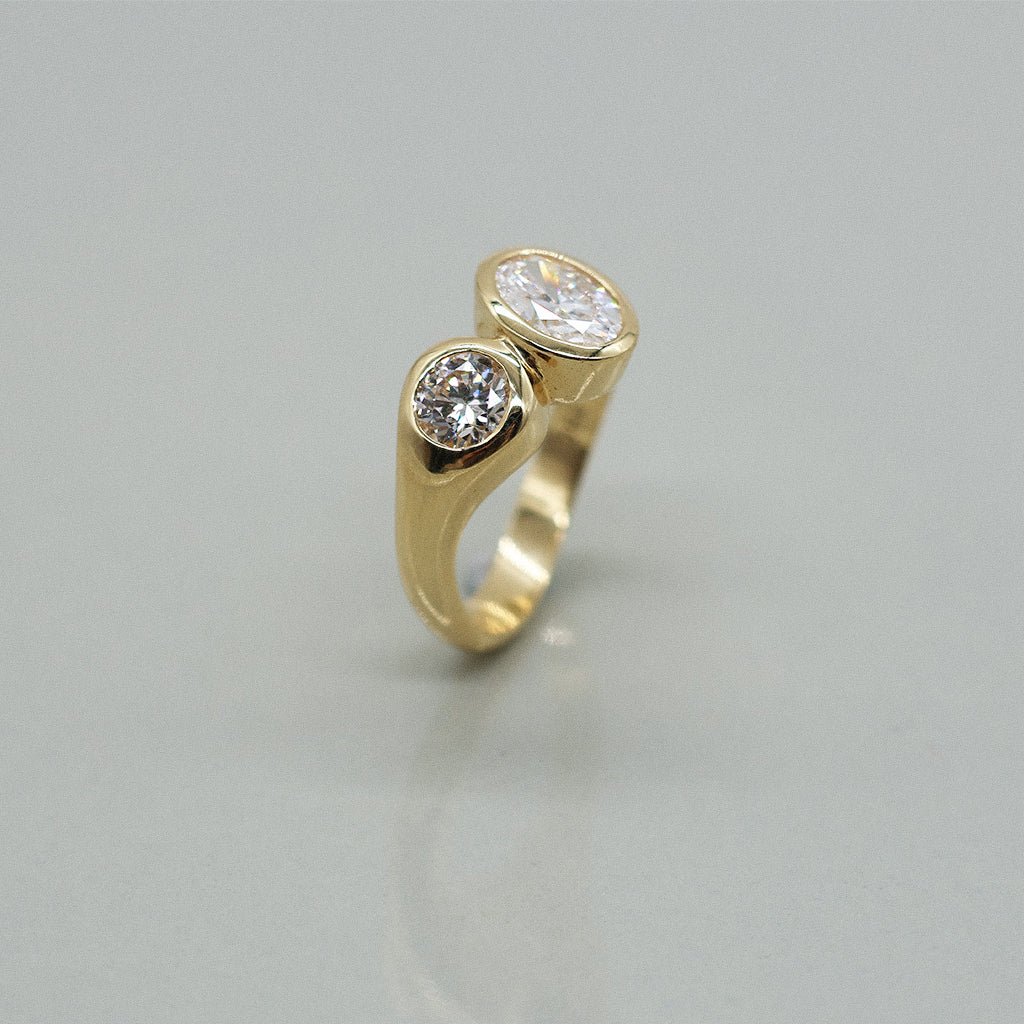 A yellow gold ring featuring a large oval brilliant-cut diamond and round diamond, set against a clean white background with a soft reflection.