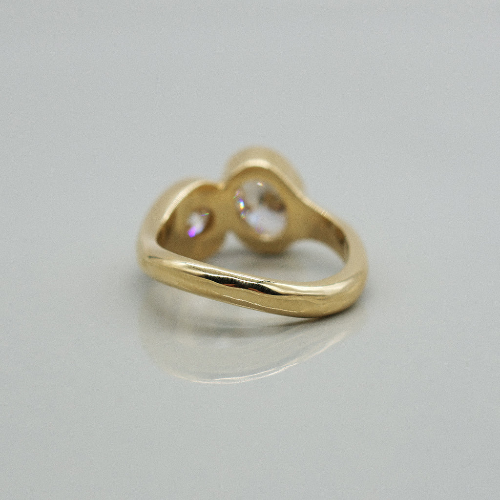 A yellow gold ring featuring a large oval brilliant-cut diamond and round diamond, set against a clean white background with a soft reflection.