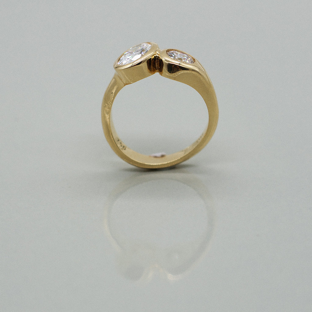 A yellow gold ring featuring a large oval brilliant-cut diamond and round diamond, set against a clean white background with a soft reflection.