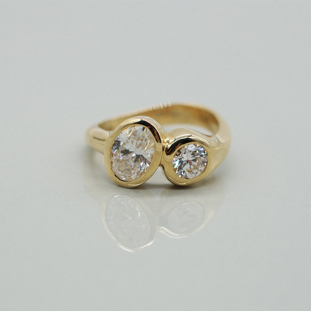 A yellow gold ring featuring a large oval brilliant-cut diamond and round diamond, set against a clean white background with a soft reflection.