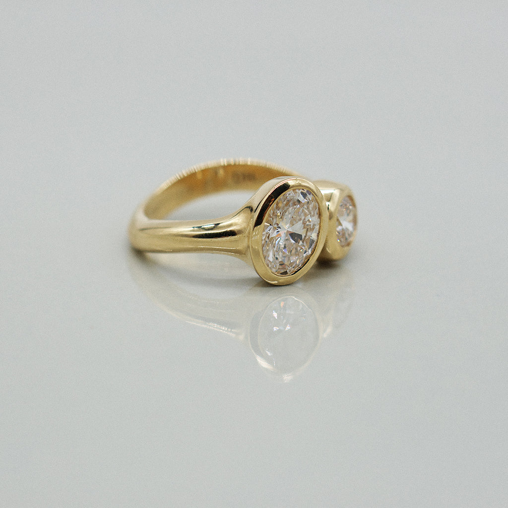 A yellow gold ring featuring a large oval brilliant-cut diamond and round diamond, set against a clean white background with a soft reflection.