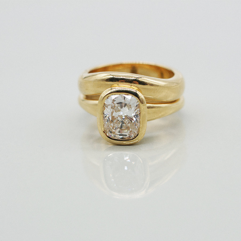A yellow gold cushion ring featuring a large cushion brilliant-cut diamond centre stone, paired with matching wedding band, set against a clean white background with a soft reflection.