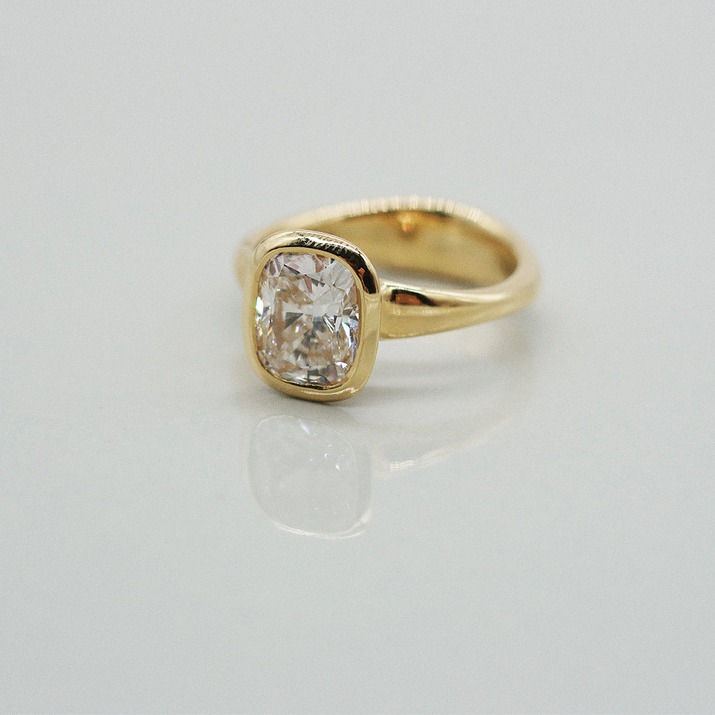 A yellow gold cushion ring featuring a large cushion brilliant-cut diamond center stone, set against a clean white background with a soft reflection.