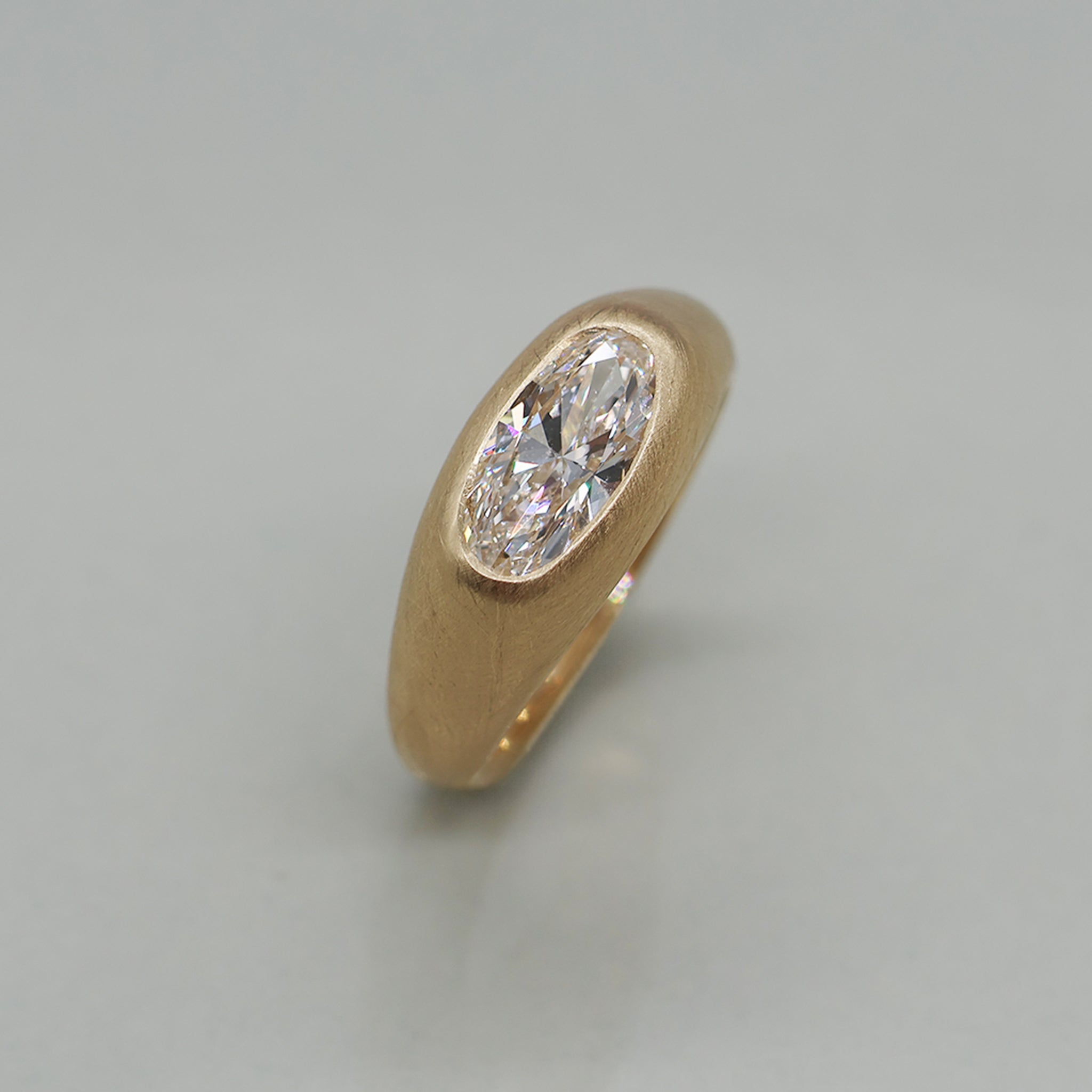 A yellow gold ring featuring a large oval brilliant-cut diamond centre stone with brushed texture, set against a clean white background with a soft reflection.