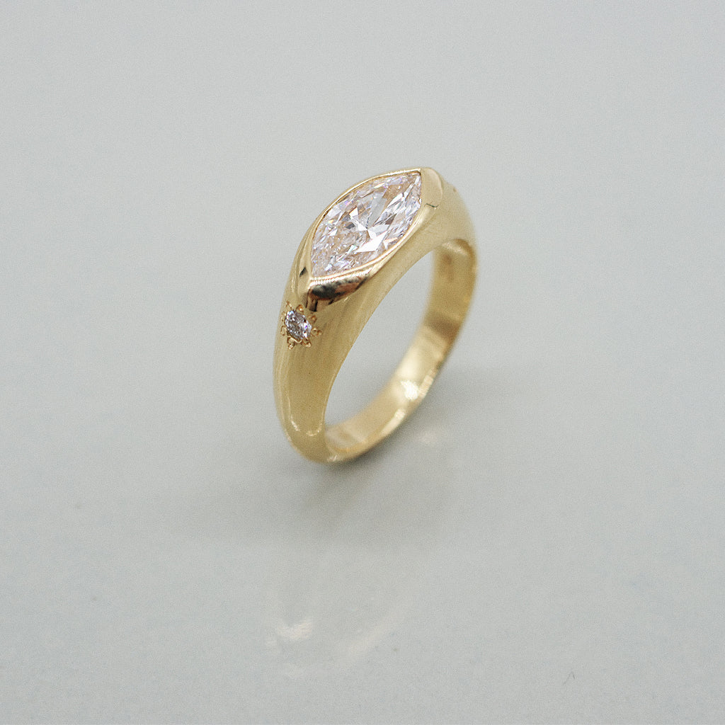 A yellow gold oval ring featuring a large marquis brilliant-cut diamond center stone and two small round diamond accents on the sides, set against a clean white background with a soft reflection.