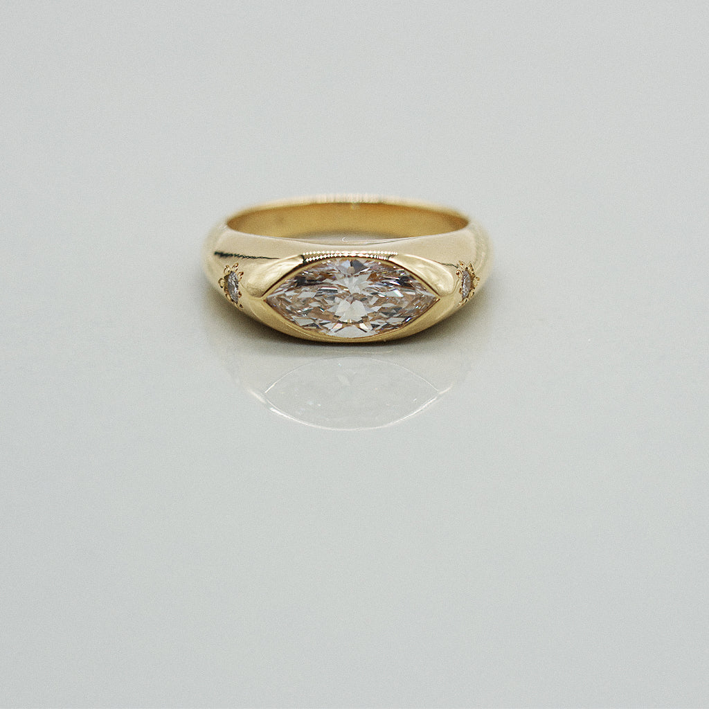 A yellow gold oval ring featuring a large marquis brilliant-cut diamond center stone and two small round diamond accents on the sides, set against a clean white background with a soft reflection.