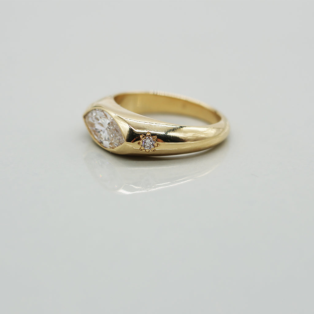 A yellow gold oval ring featuring a large marquis brilliant-cut diamond center stone and two small round diamond accents on the sides, set against a clean white background with a soft reflection.