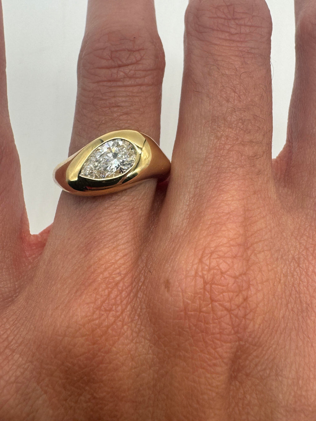 A pear-shaped white diamond set in a gold ring with a wavy signet band, displayed on a person's finger.