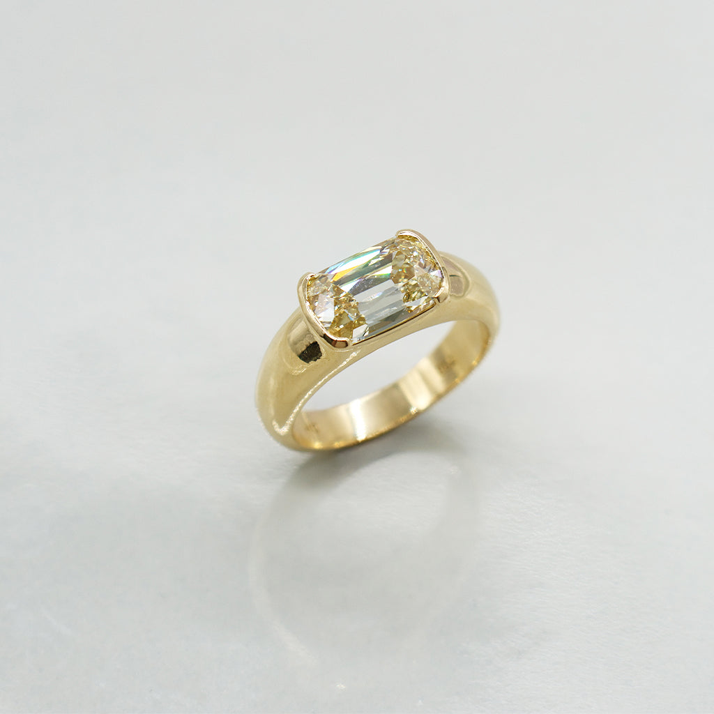 A yellow gold ring featuring a rectangular emerald-cut iridescent gemstone, set in a four-prong setting on a light gray reflective surface.