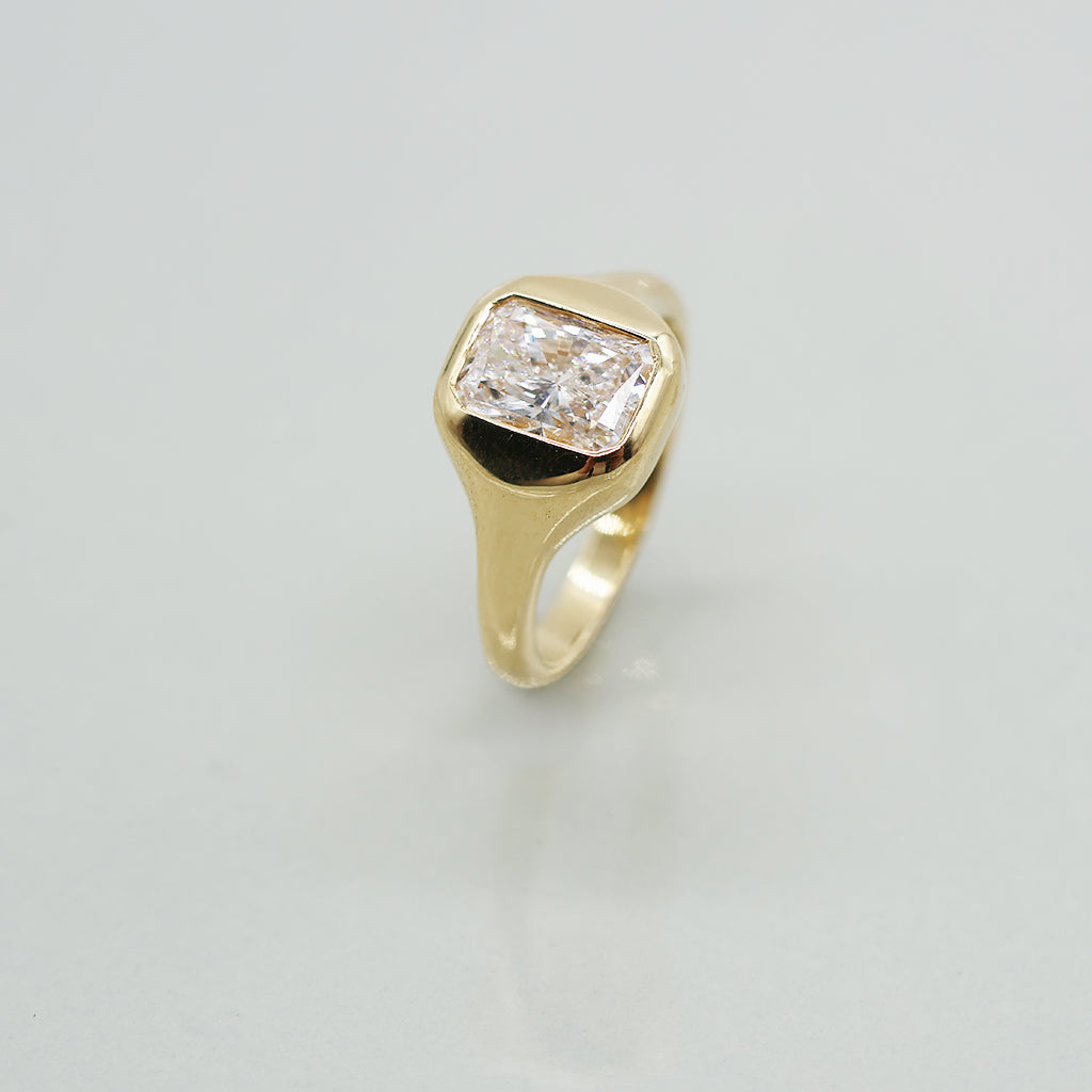 A yellow gold ring with a radiant cut white diamond in a bezel setting, featuring a wavy, layered gold band, photographed on a light gray reflective surface.