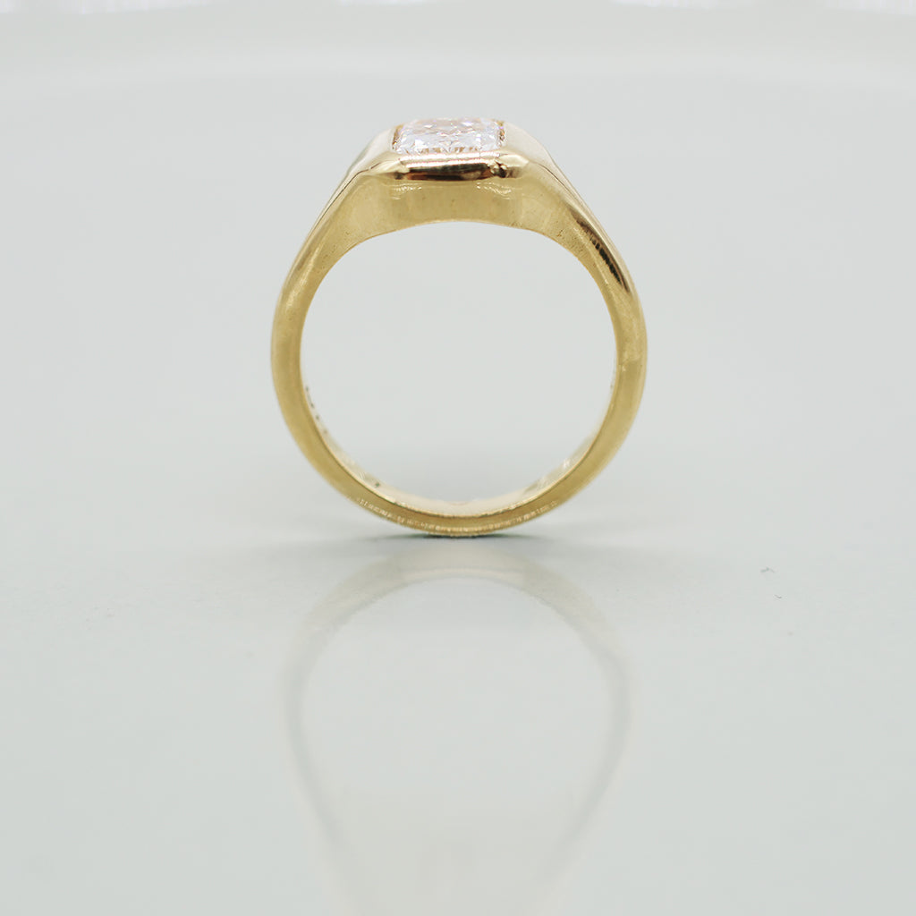 A yellow gold ring with a radiant cut white diamond in a bezel setting, featuring a wavy, layered gold band, photographed on a light gray reflective surface.