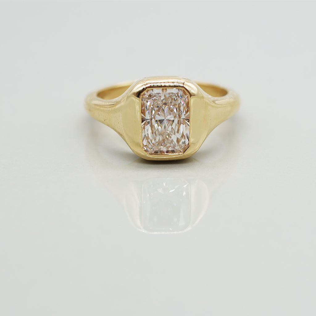 A yellow gold ring with a radiant cut white diamond in a bezel setting, featuring a wavy, layered gold band, photographed on a light gray reflective surface.