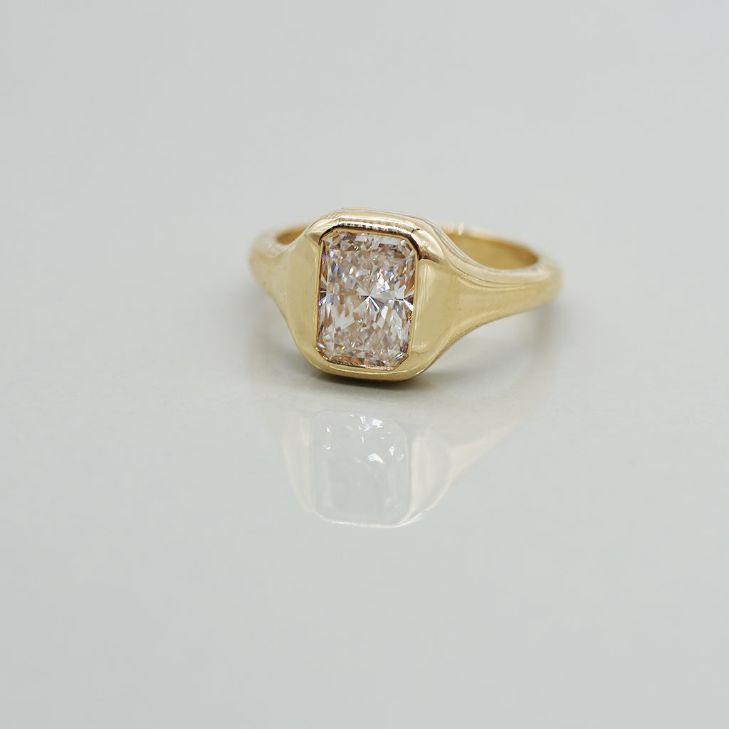 A yellow gold ring with a radiant cut white diamond in a bezel setting, featuring a wavy, layered gold band, photographed on a light gray reflective surface.