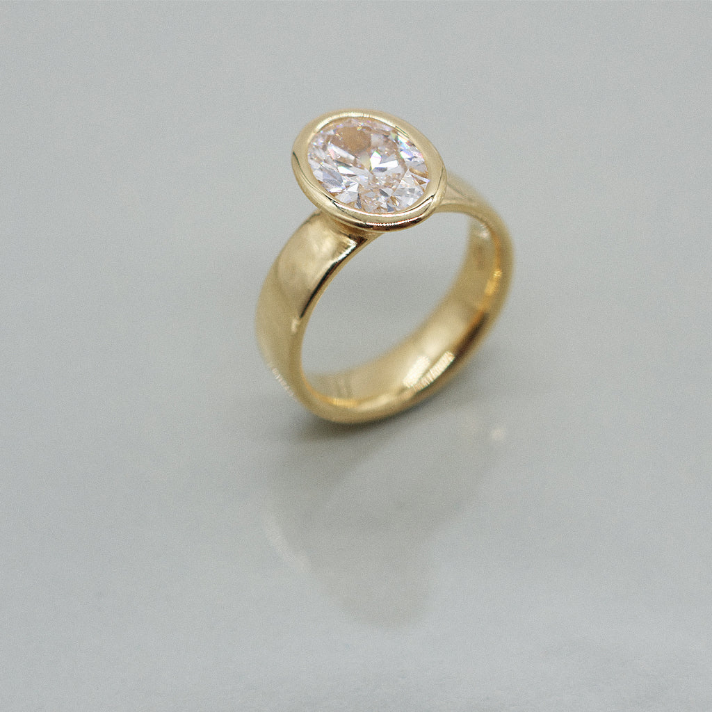 Gold ring with oval diamond centre on a wide ring band. Set against a clean white background with a soft reflection.