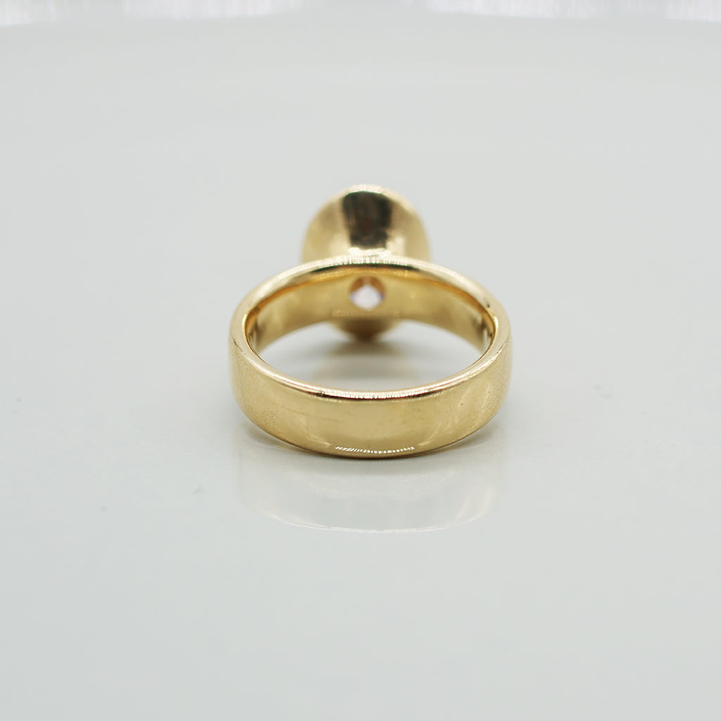 Gold ring with oval diamond centre on a wide ring band. Set against a clean white background with a soft reflection.