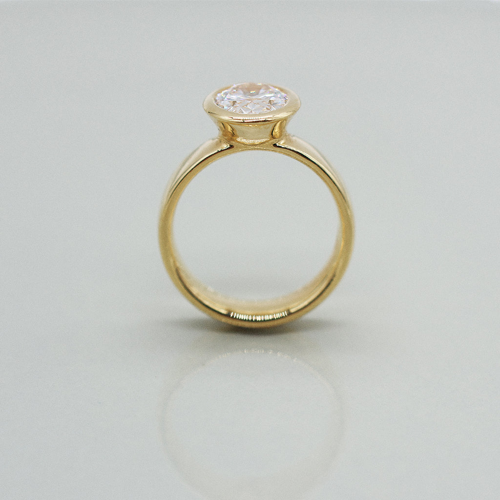 Gold ring with oval diamond centre on a wide ring band. Set against a clean white background with a soft reflection.