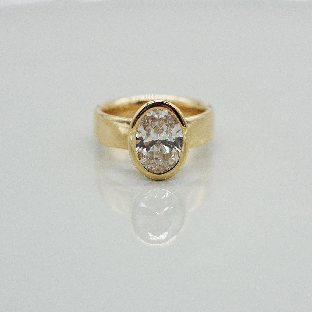Gold ring with oval diamond centre on a wide ring band. Set against a clean white background with a soft reflection.
