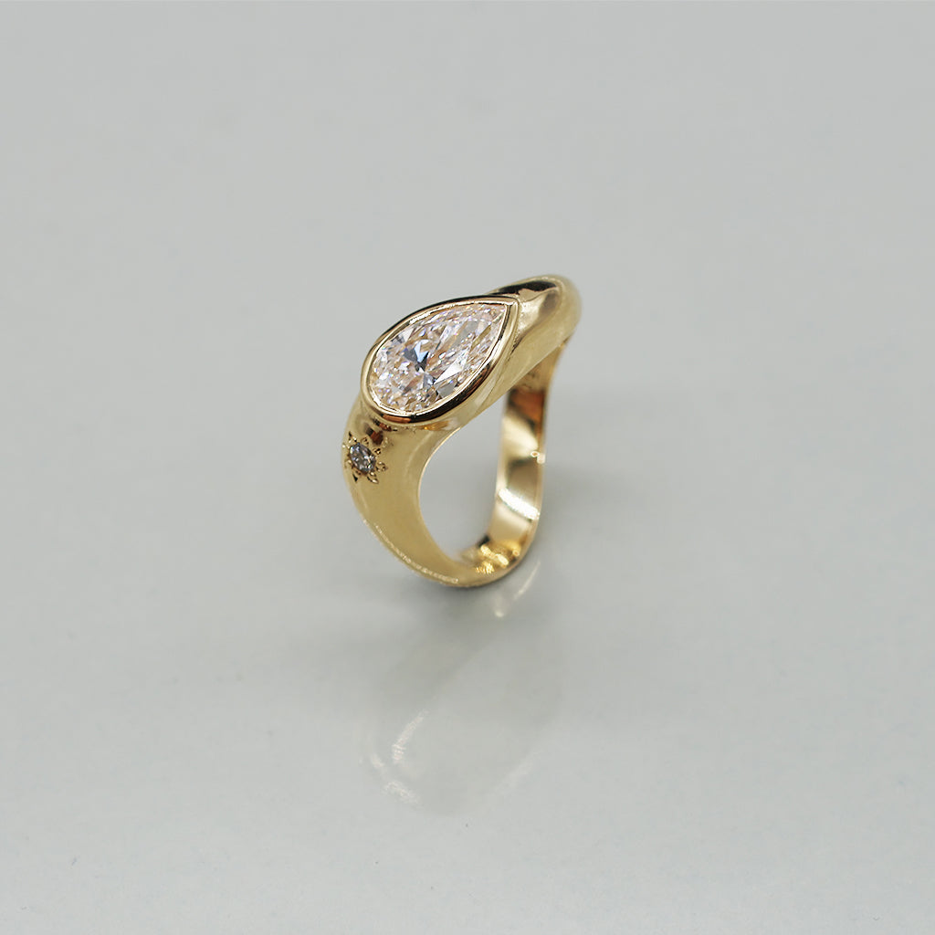 A yellow gold oval ring featuring a large pear brilliant-cut diamond centre stone and a small round stone on a wavy band, set against a clean white background with a soft reflection.
