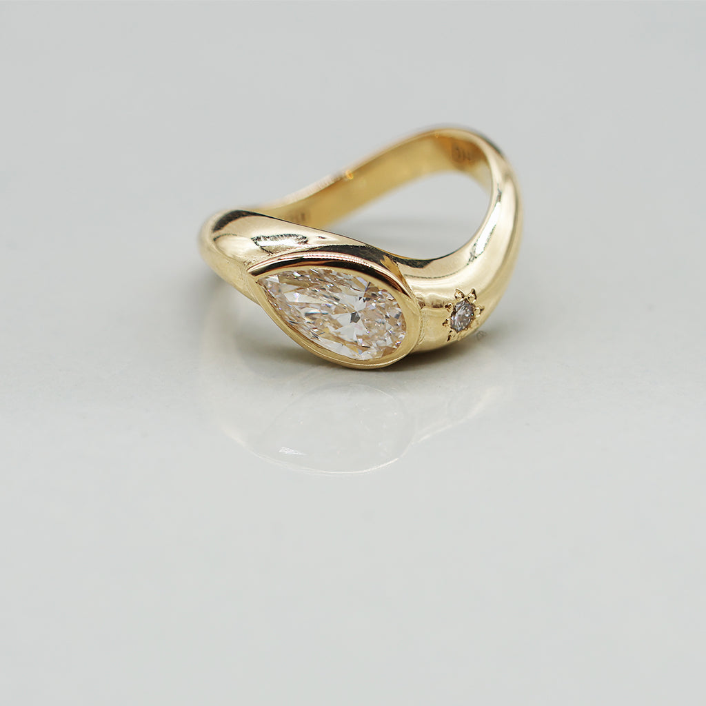 A yellow gold oval ring featuring a large pear brilliant-cut diamond centre stone and a small round stone on a wavy band, set against a clean white background with a soft reflection.
