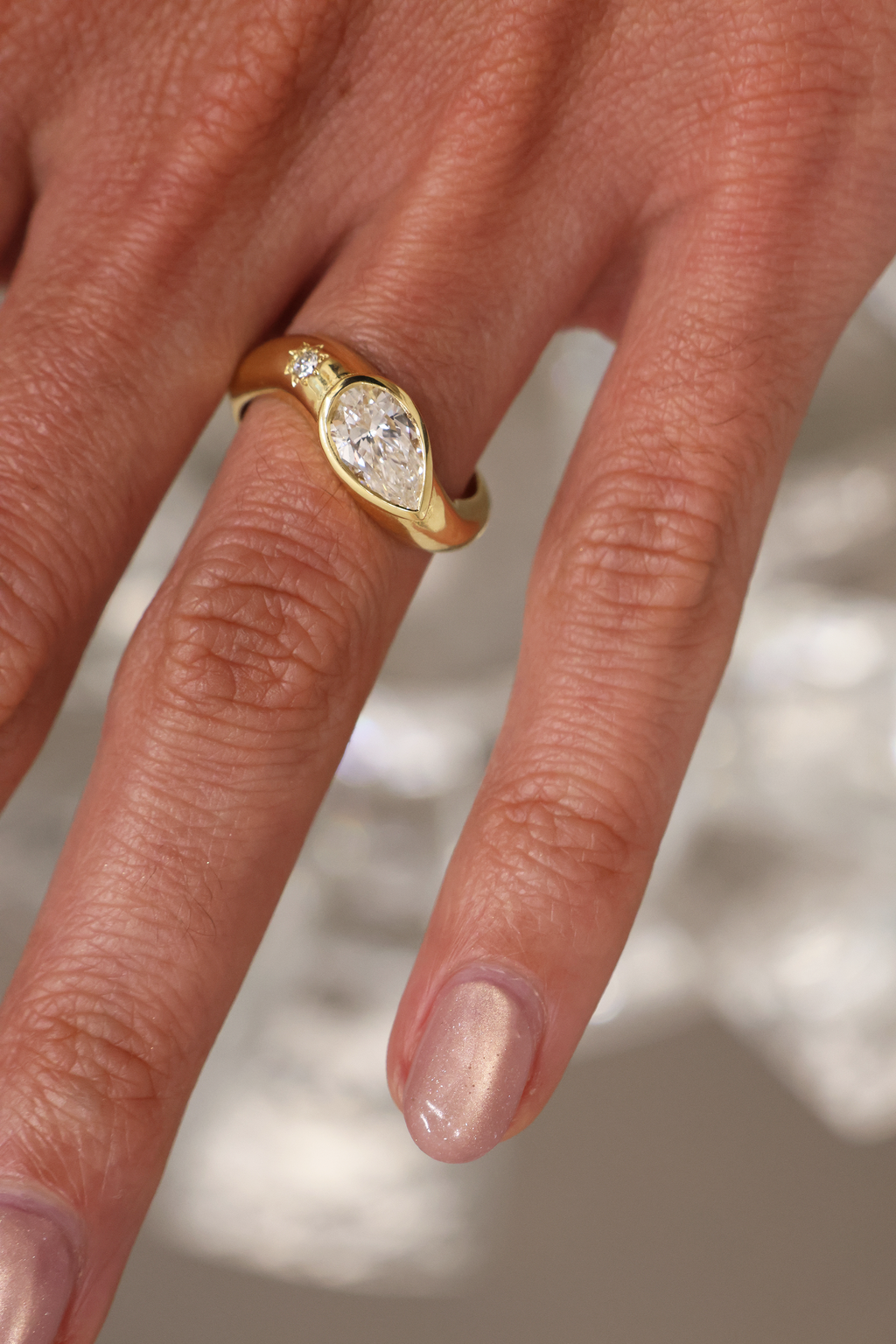 A yellow gold oval ring featuring a large pear brilliant-cut diamond centre stone and a small round stone on a wavy band, on the hand set against a neutral blurred background.