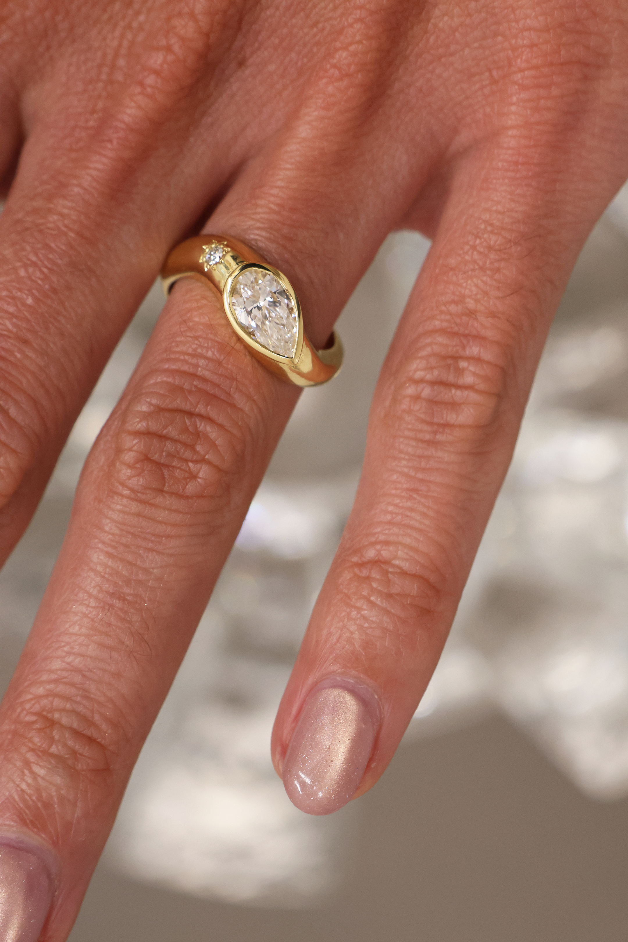 A yellow gold oval ring featuring a large pear brilliant-cut diamond centre stone and a small round stone on a wavy band, on the hand set against a neutral blurred background.