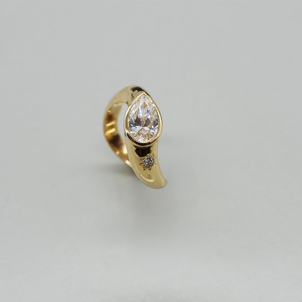 A yellow gold oval ring featuring a large pear brilliant-cut diamond centre stone and a small round stone on a wavy band, set against a clean white background with a soft reflection.