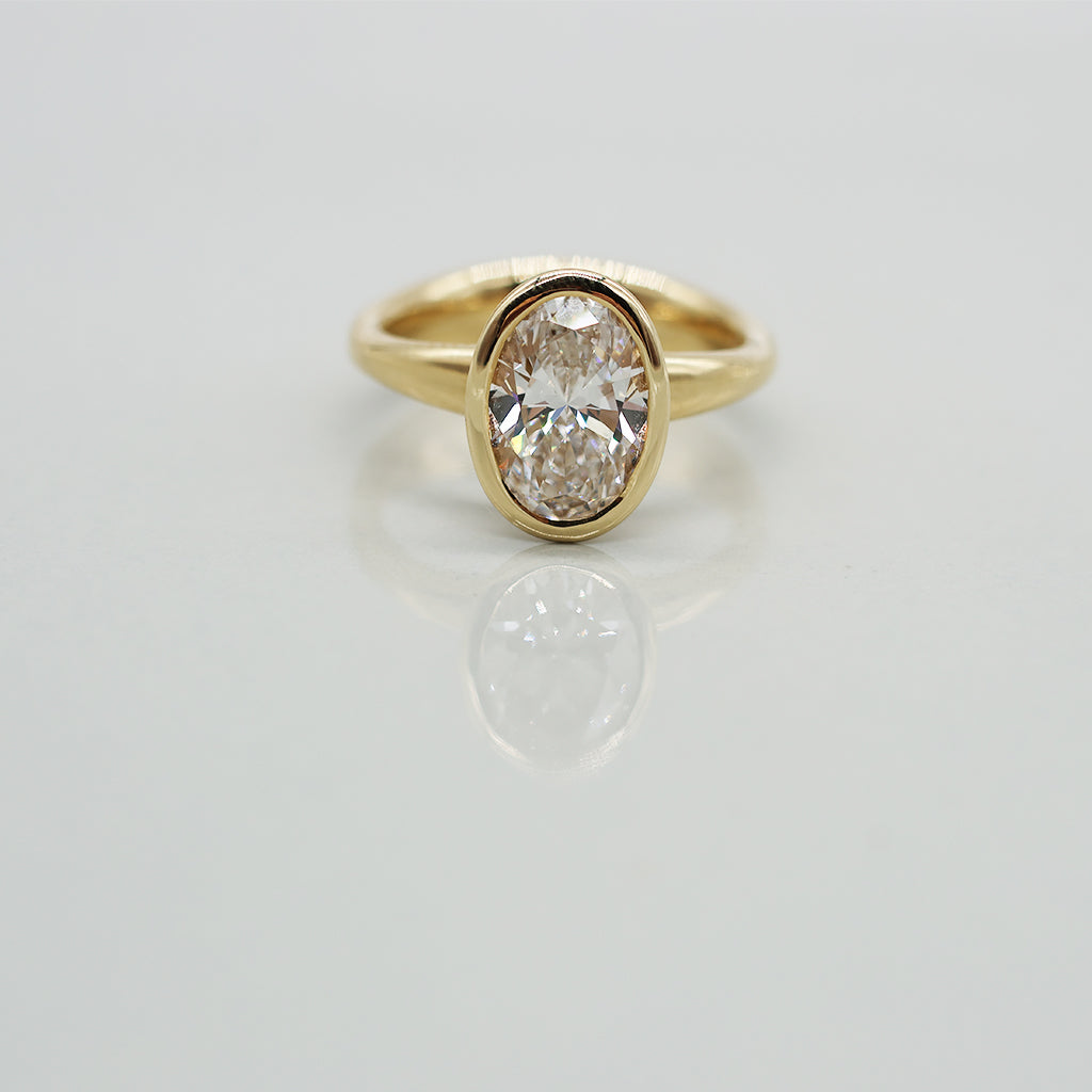Gold ring with an oval diamond set in full bezel on a light gray background
