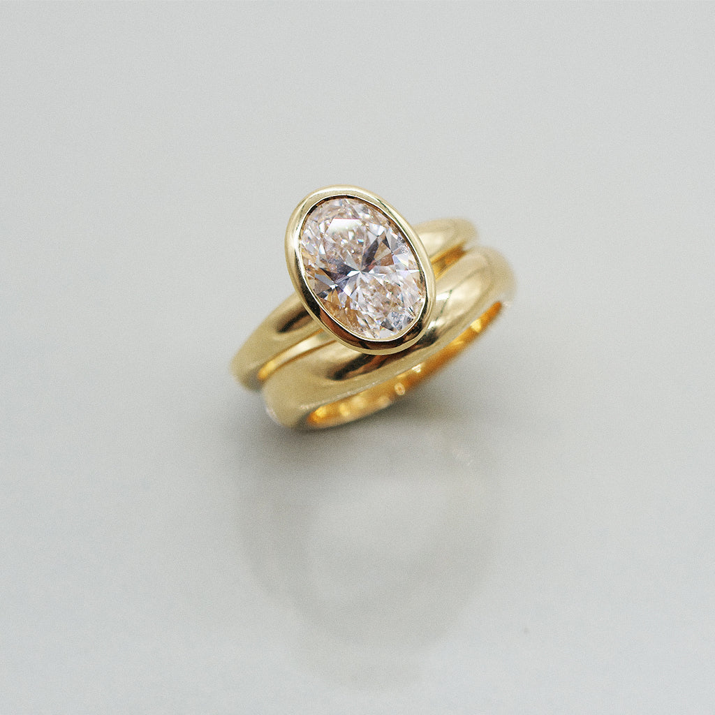 Gold ring with an oval diamond set in full bezel paired with matching wedding band on a light gray background