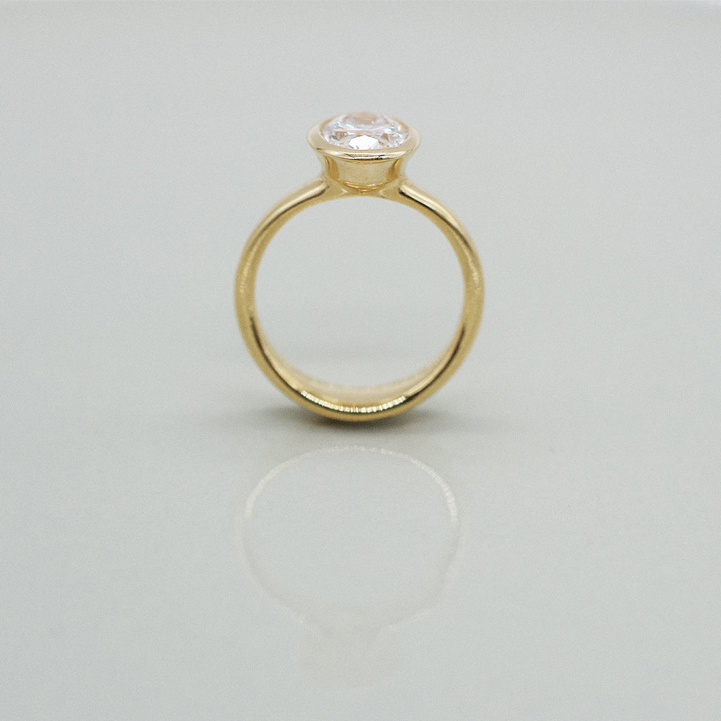Gold ring with an oval diamond set in full bezel on a light gray background